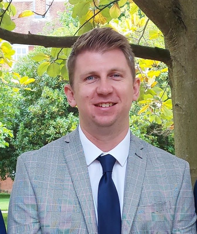 A white man in a light grey suit and navy tie smiles at the camera with trees and foliage in the background