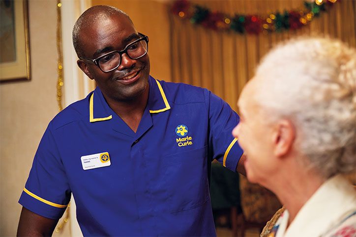 Marie Curie Nurse Isaac smiles as he speaks to a woman. He's wearing a blue Marie Curie uniform.