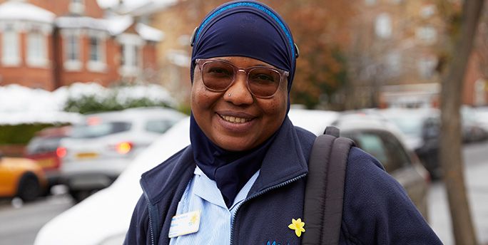 A Marie Curie Nurse stands outside of a house. She smiles as she poses for a photograph. She's wearing sunglasses and a blue Marie Curie uniform.