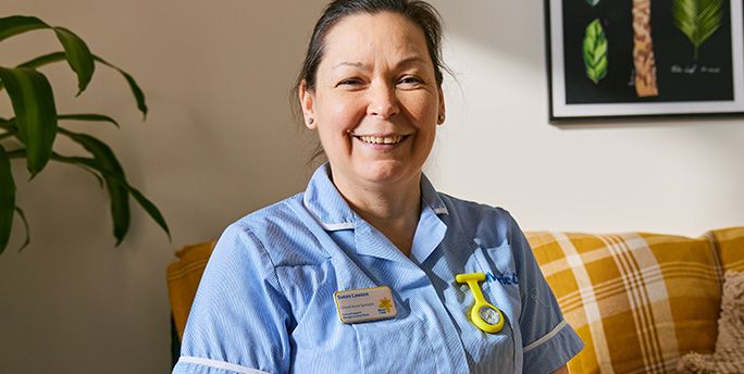 Marie Curie Nurse Susan sits on a yellow sofa and smiles as she poses for a photograph.