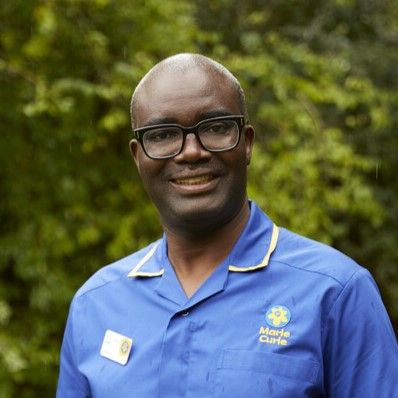 Marie Curie Nurse Isaac smiles as he poses for a photograph outside near trees. He's wearing a blue Marie Curie uniform and black glasses. 