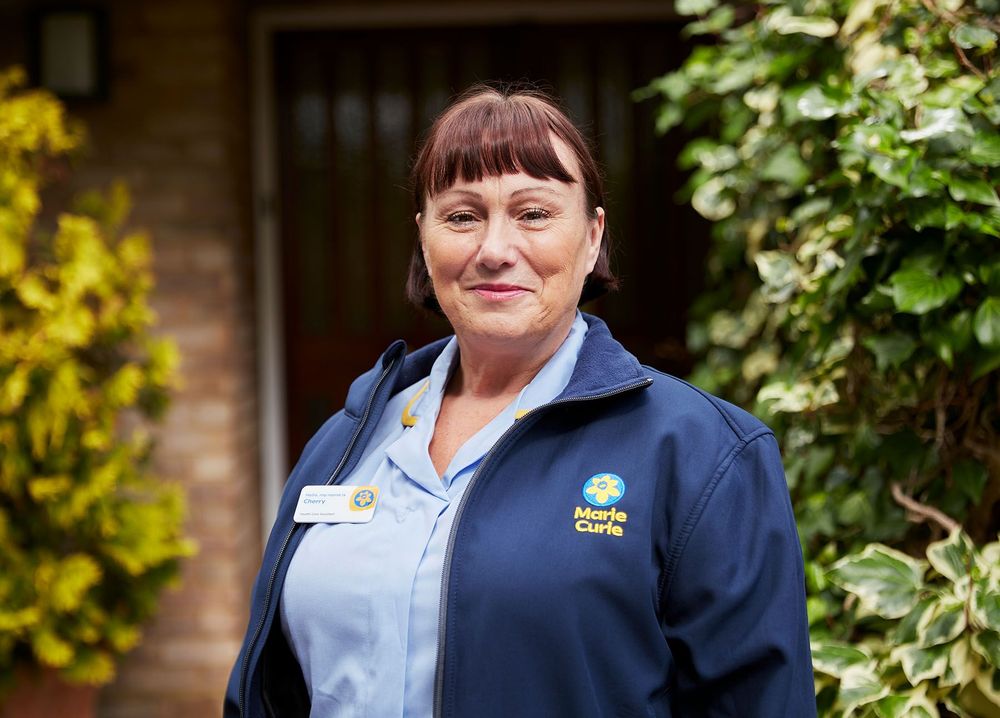 A Marie Curie Nurse smiles and poses for a photograph in front of a door and outdoor plants. 