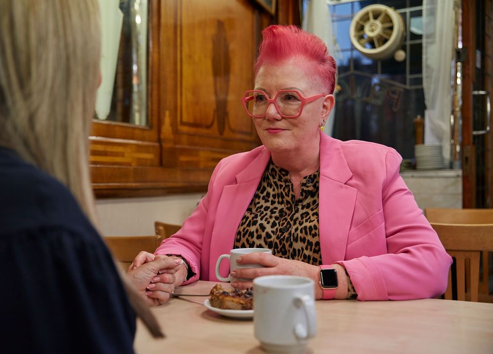 A woman with bright pink hair is sat at a table with a cup of tea in her hand as she speaks to a woman in front of her.