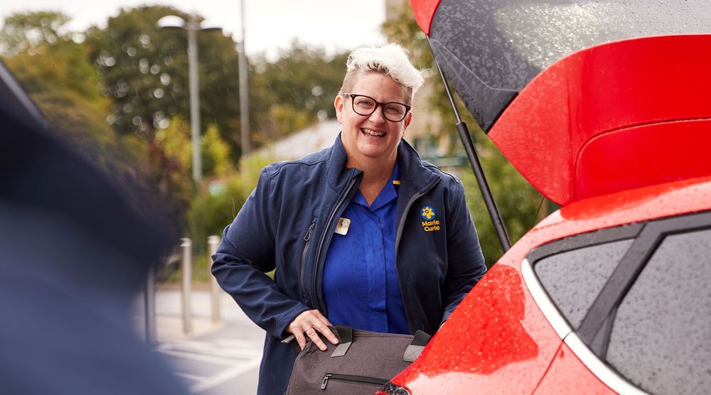 A Marie Curie Nurse puts a bag in the boot of a red car while she smiles