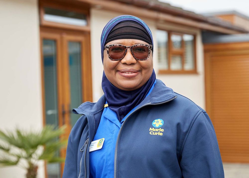 A Marie Curie Nurse stands outside of a house. She smiles as she poses for a photograph. She's wearing sunglasses and a blue Marie Curie uniform.