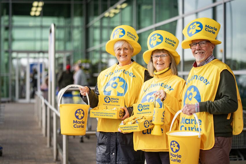 Three Marie Curie volunteers wearing yellow tabards and hats hold collection buckets, tins and a box of daffodil pins as they stand in a row outside a supermarket