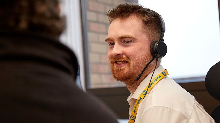 A man with a headset smiles. He's sat a desk and wearing a yellow Marie Curie lanyard.