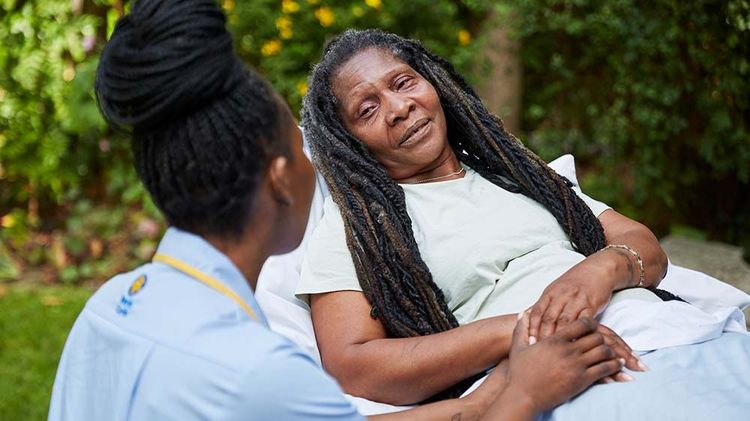 A Marie Curie Nurse consoles a patient sitting in a hospice bed. They are outside in the hospice gardens.