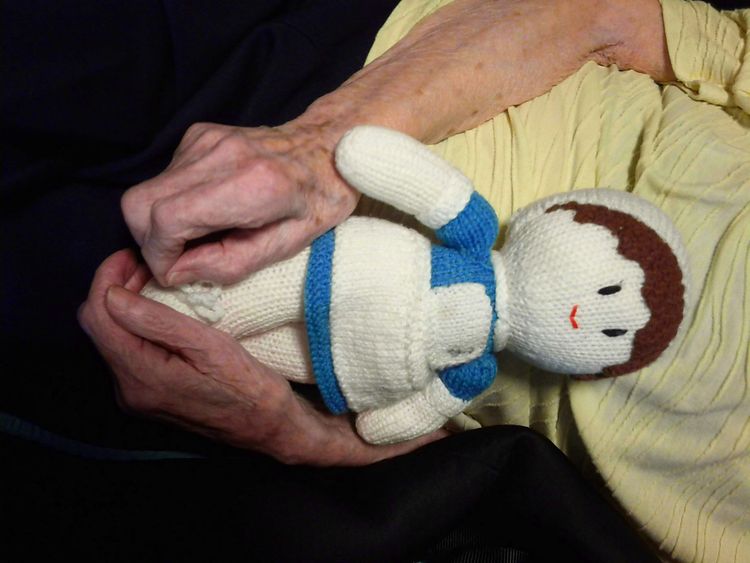 A knitted doll with a blue dress nestles in the lap of an elderly woman