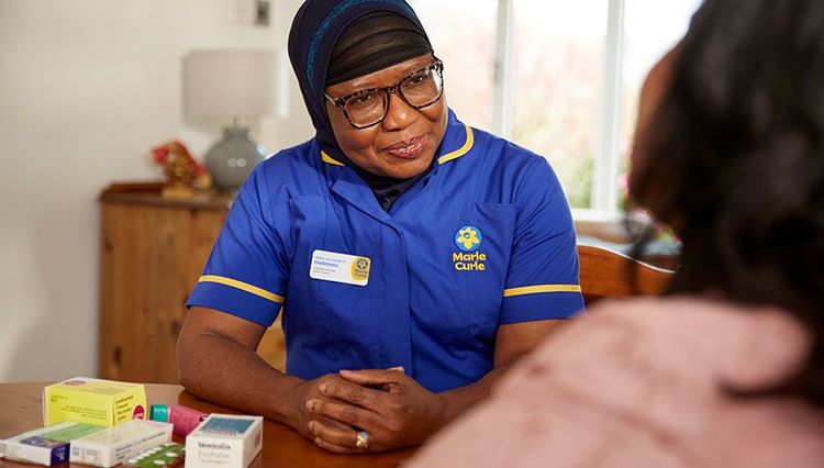 A Marie Curie nurse sits at a table and speaks to a patient. Multiple medications are visible on the dining room table.