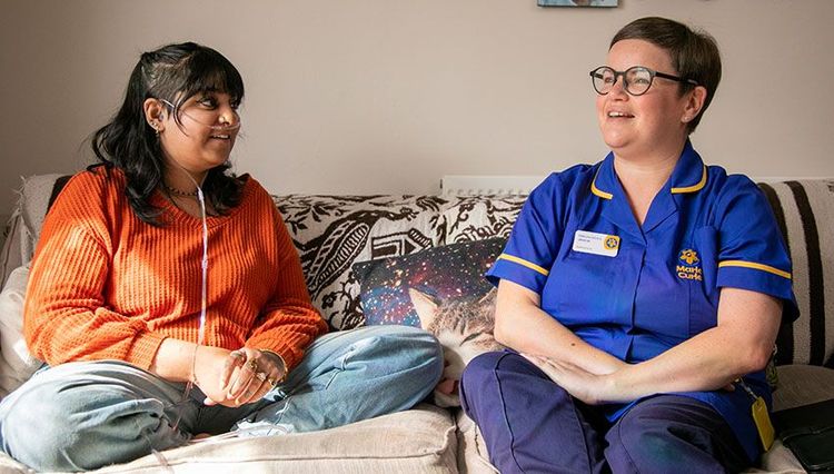 A Marie Curie Nurse sits next to a patient on a living room sofa.