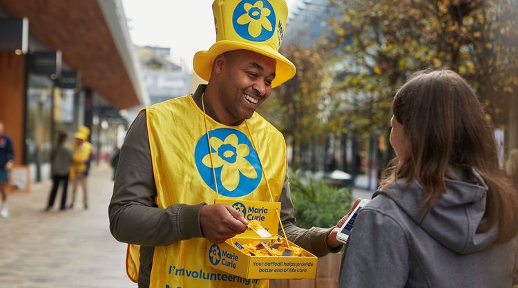 Marie Curie collector wearing a yellow top hat and yellow vest hands out a Marie Curie daffodil pin to a woman on the street