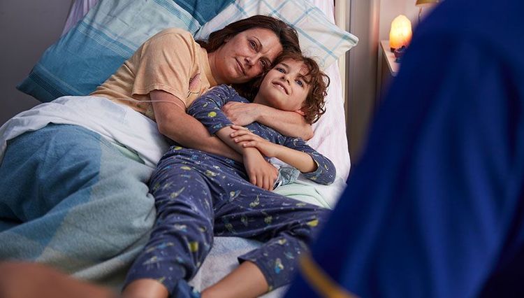 A mother and child hug as they next to each other in a hospice bed.