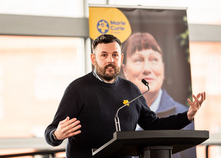 A speaker stands at a podium giving a speech. They're wearing a daffodil pin on their black sweatshirt.