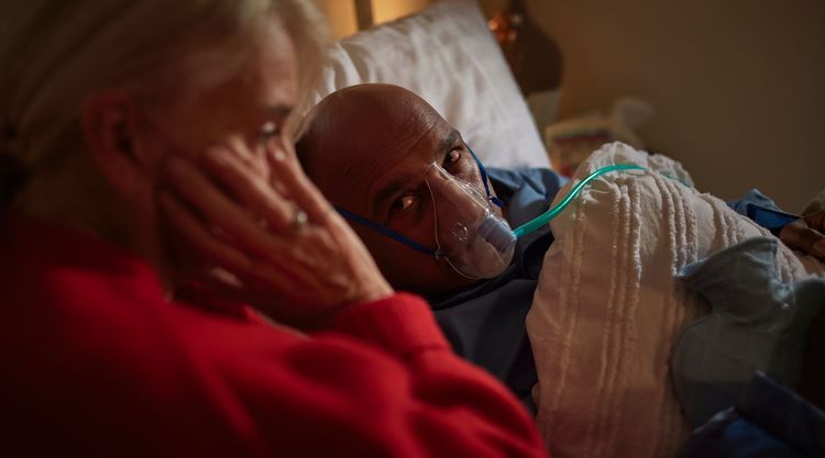 A man lies in bed with an oxygen mask on while a worried women sits next to him, her hand on her cheek