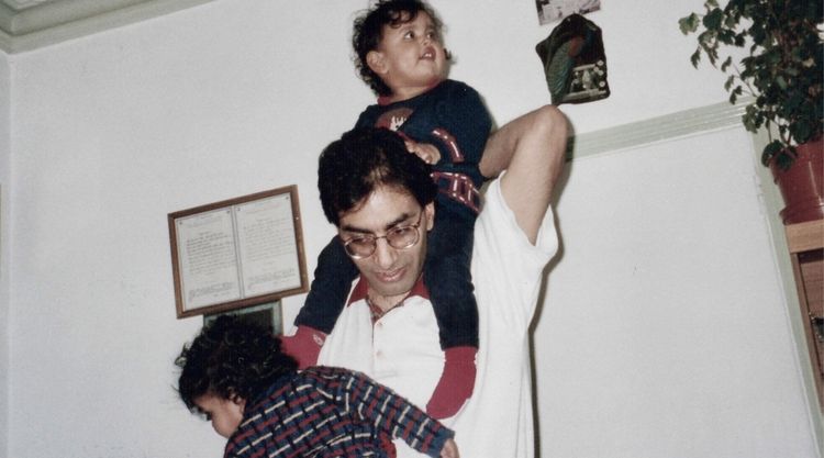 A younger Harmi holding one baby daughter in one arm while he supports his other baby daughter with his other arm as she sits on his shoulders