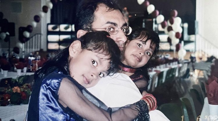 A younger Harmi sitting at a party table holding one daughter as a little girl with the other daughter hugging his shoulder.