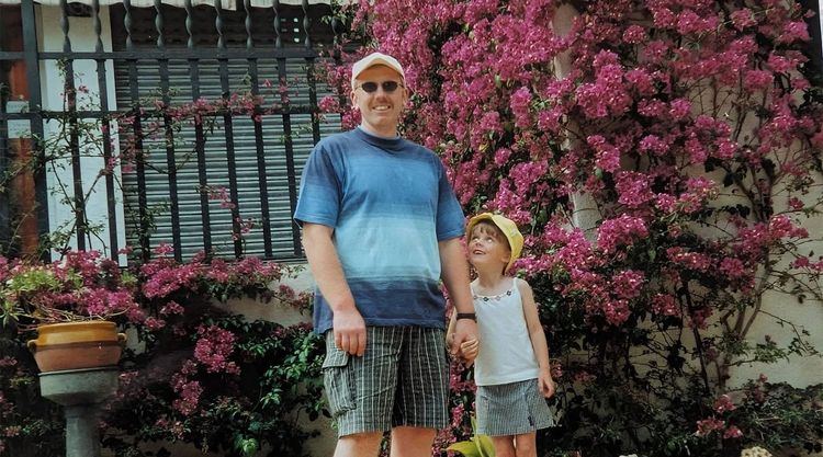 A smiling father and his young daughter hold hands in front of a house covered with a pink flowering vine