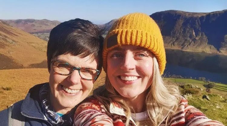Two woman, one wearing a yellow hat, smile in a selfie taken in front of a mountain and lake