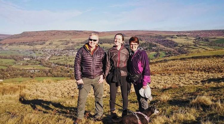 A man and two women in walking gear smile at the camera standing on open moorland on a sunny day