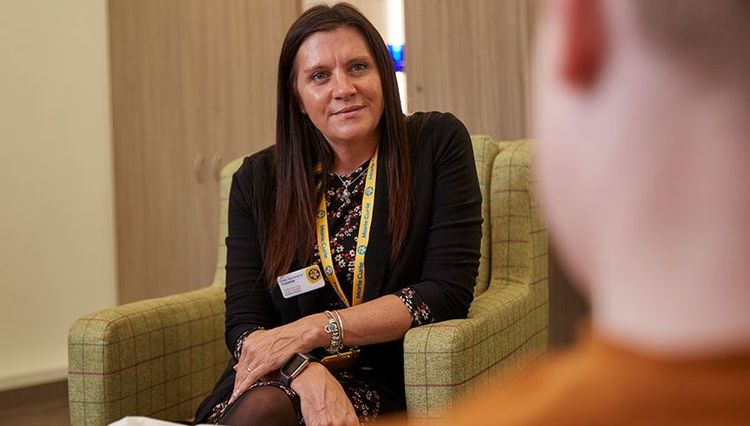 A woman sits in a chair while in conversation with a man. She's wearing a yellow Marie Curie lanyard and name badge.