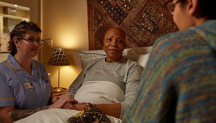 A patient sits up in her bed and a Marie Curie Nurse is seated beside her, holding her hand. 